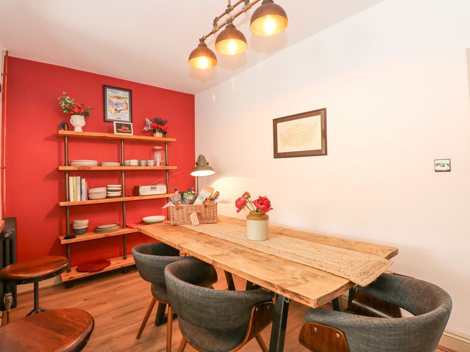 A dining area with a wooden table, grey upholstered chairs, a red wall, and a bookshelf with various items.