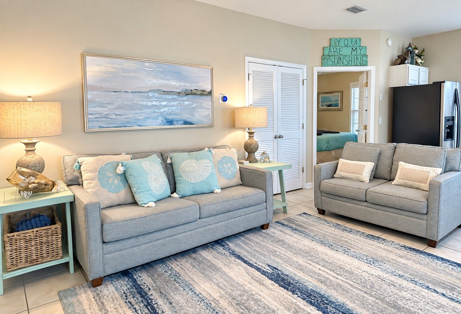 A living room with a grey sofa, a matching armchair, a painting of a beach scene, and a blue and white striped rug.