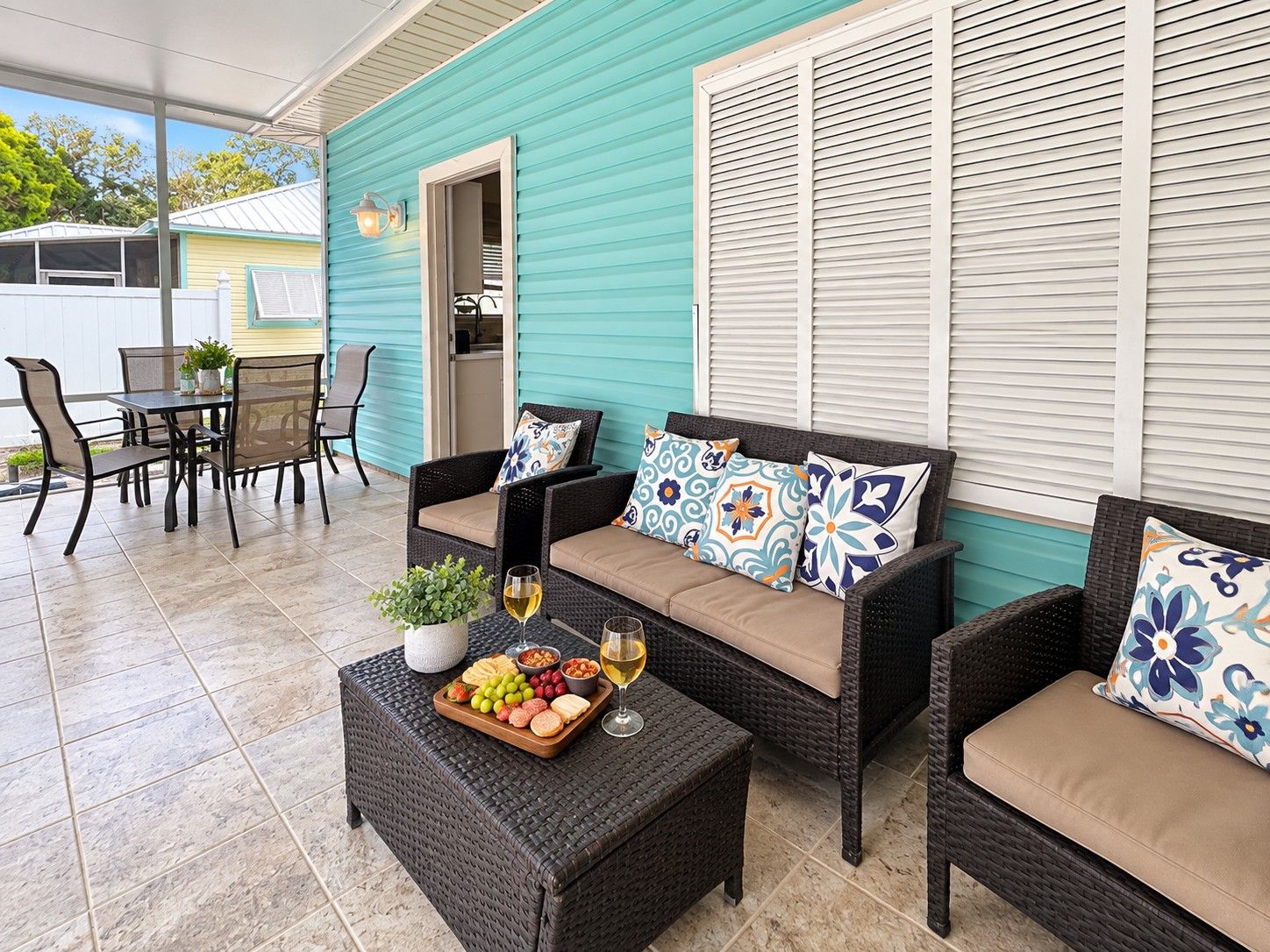 Un espacio de patio cubierto con mobiliario de mimbre, una mesa con una bandeja de frutas y copas de vino, y un edificio color turquesa al fondo.