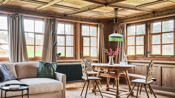 A cozy room with a wooden ceiling, large windows, a sofa, a dining table with chairs, and a radiator.