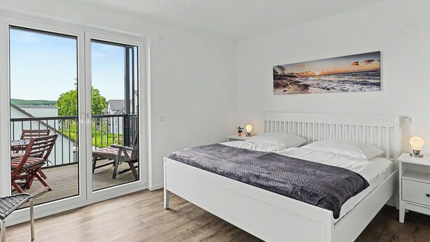 Een slaapkamer met een bed, nachtkastjes, een stoel, een balkon met uitzicht en een schilderij aan de muur.