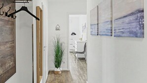 Een gang met een houten wand, een kapstok, een kamerplant, een witte deur en een bureau met een stoel.
