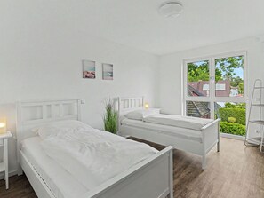 Une chambre avec deux lits, une table de chevet, une plante et une fenêtre donnant sur un jardin et des bâtiments.