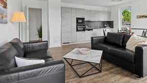 Een moderne woonkamer met twee zwarte leren banken, een glazen salontafel en een keuken met grijze kasten en apparatuur.