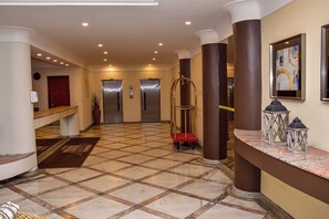 Eine Hotelhalle mit Check-in-Schalter, Rezeption und einer Treppe sowie zwei Aufzügen.