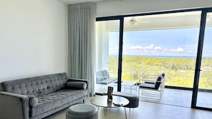 A modern living room with a grey sofa, a round coffee table, and two ottomans. There is a glass dining table with chairs, and a large sliding glass door leading to a balcony with a view of greenery and mountains. - Chic 2BR Apt 1km from Playa Bonita (Las Terrenas)