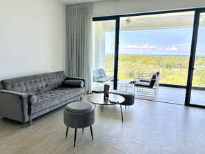 Ein modernes Wohnzimmer mit einem grauen Sofa, einem runden Couchtisch und zwei Poufs. Es gibt einen Glastisch mit Stühlen und eine große Schiebetür, die auf einen Balkon mit Blick auf Grünflächen und Berge führt.