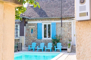 A stone building with a swimming pool, blue shutters, and four blue chairs.