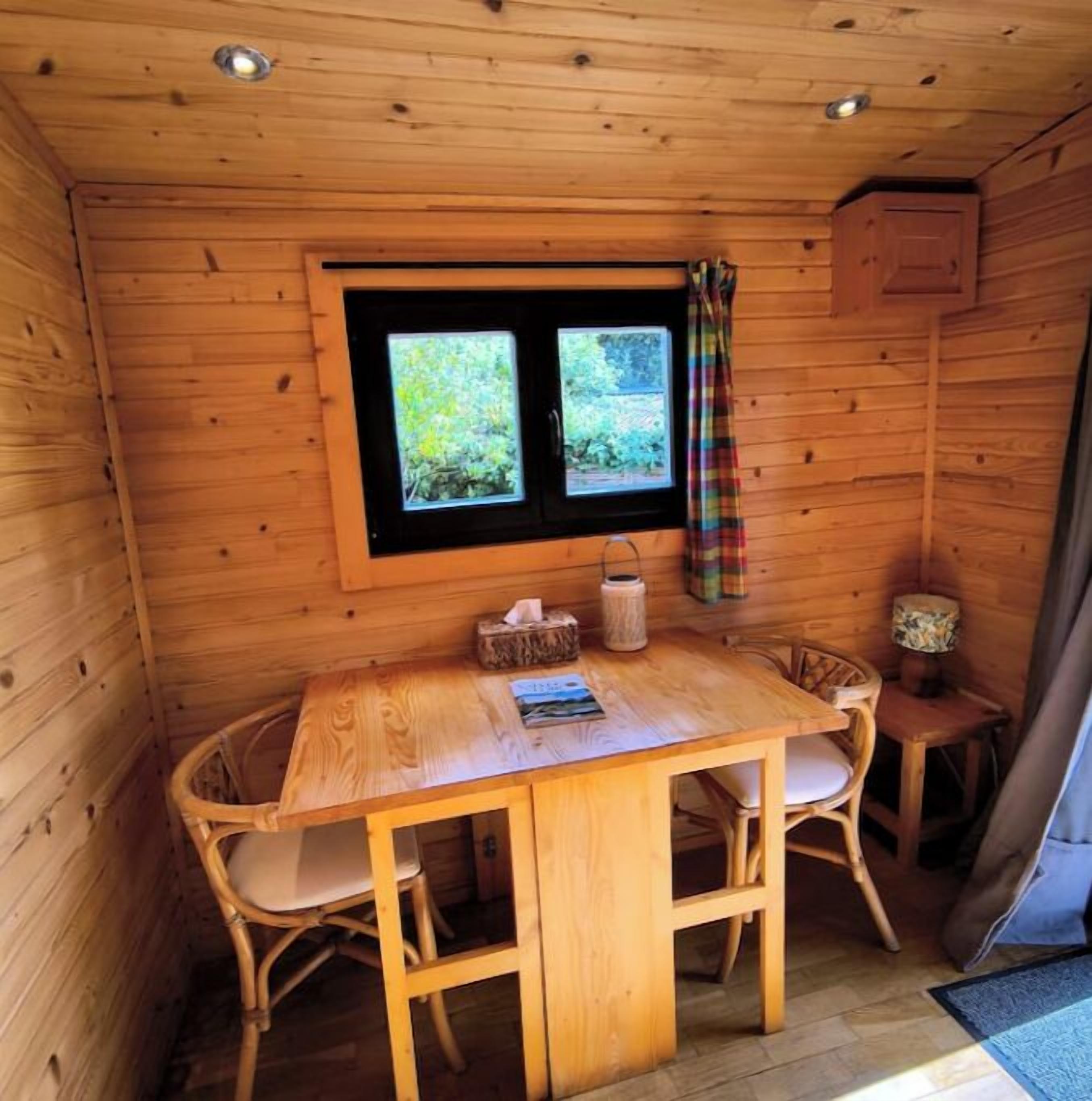 Eine Holzhütte mit Esstisch und Stühlen, ein Fenster mit Blick auf Grünflächen und ein karierter Vorhang.