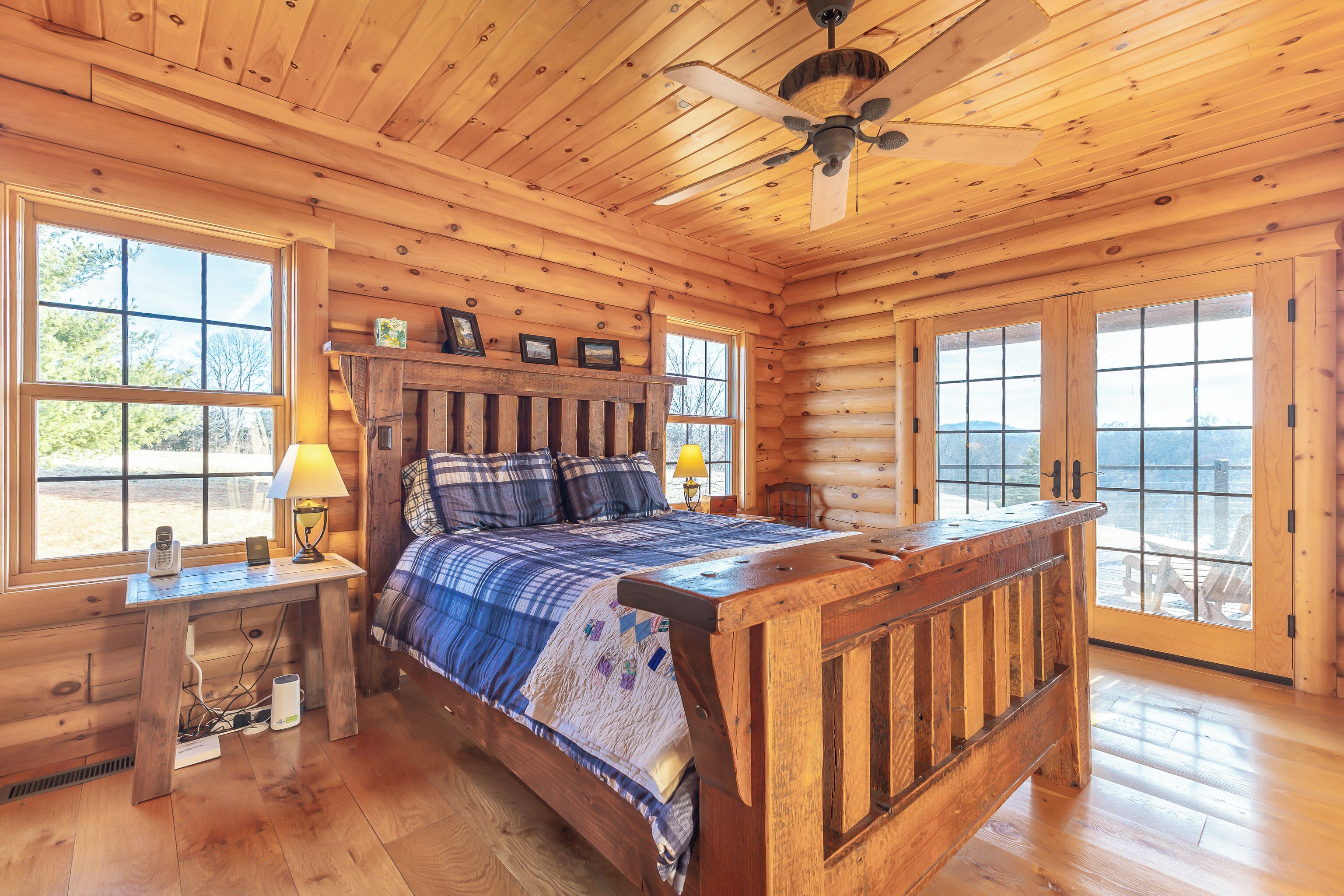 A log cabin bedroom with a wooden bed, a nightstand, a lamp, and a window with a view of the outdoors.