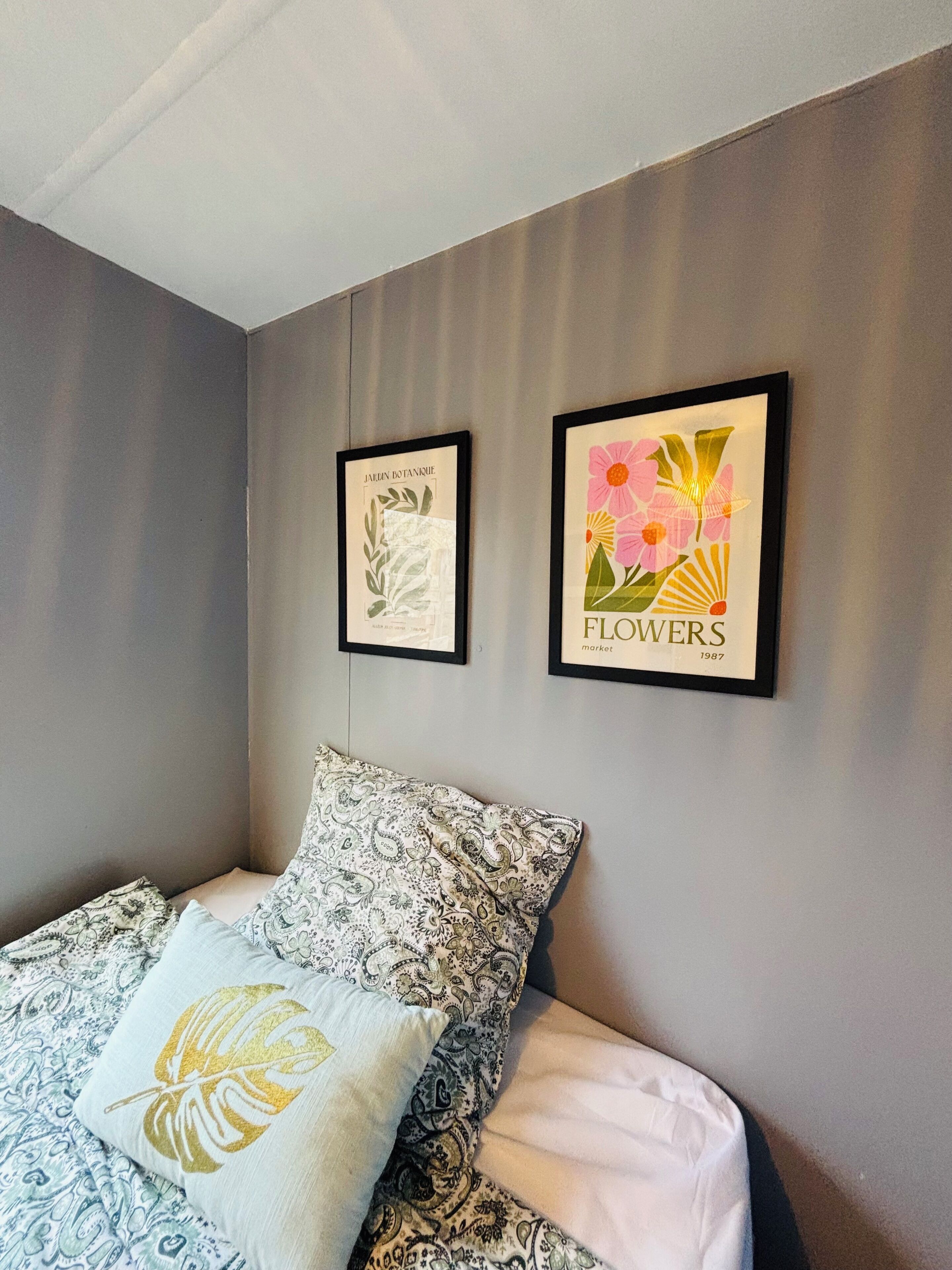A bedroom with a bed, two framed botanical prints, and a patterned pillow.