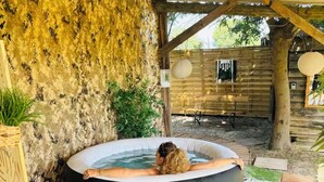 Un bain à remous sous une pergola en bois, adossé à un mur rustique, entouré d’une terrasse en bois et de plantes en pot.