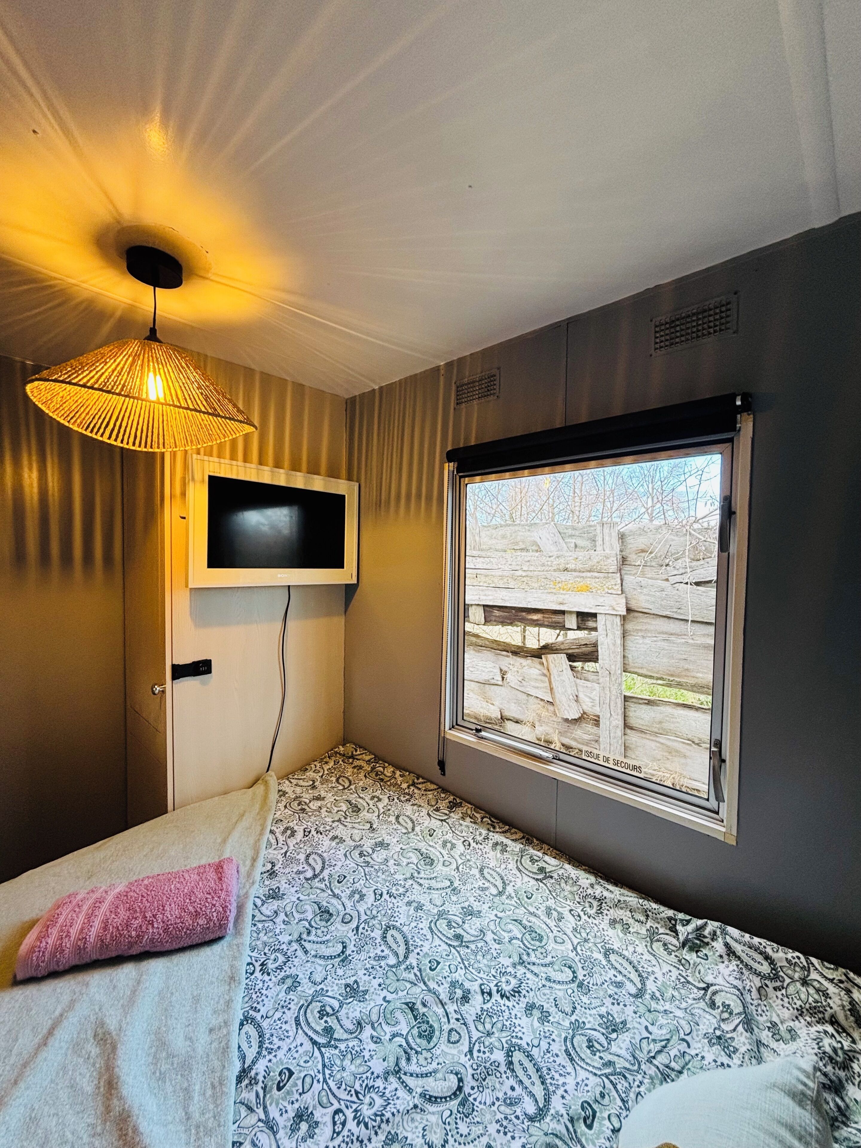 Une chambre avec un lit, un téléviseur fixé au mur, une fenêtre donnant sur une clôture en bois et un luminaire suspendu.