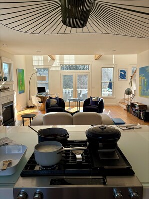 A modern kitchen with a gas stove, pots, and a kettle. There is a dining area with a sofa and chairs, a fireplace, and artwork on the walls. - August or weekly rental bike to beach, walk to tennis! (Westerly)