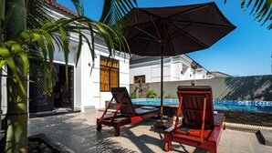 A poolside area with two wooden lounge chairs and a large umbrella, a stone-lined water feature, and a white building with decorative doors. - 2Bdrm with Salt Water Pool 15 mins drive from Center (Krong Siem Reap)