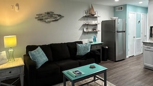 Een woonkamer met een zwarte bank, een zeegroene salontafel, een bureau met planken, een plafondventilator en een koelkast.