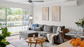 A living room with a grey sofa, a round wooden coffee table, and a dining area with a glass table set for two. - Kangaroo Sanctuary (Sanctuary Point)