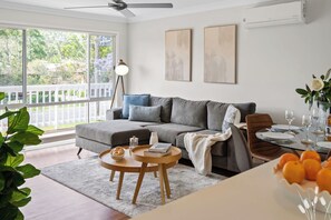 A living room with a grey sofa, a round wooden coffee table, and a dining area with a glass table set for two. - Kangaroo Sanctuary (Sanctuary Point)