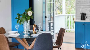 A dining table set with plates, glasses, and a bottle, surrounded by chairs, with a view of a blue exterior and a kitchen area. - Kangaroo Sanctuary (Sanctuary Point)