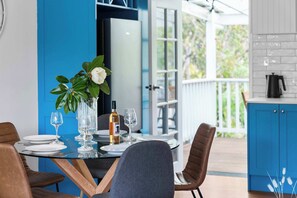 A dining table set with plates, glasses, and a bottle, surrounded by chairs, with a view of a blue exterior and a kitchen area. - Kangaroo Sanctuary (Sanctuary Point)