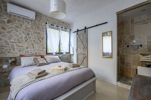 Ein Schlafzimmer mit Wand im Steinoptik, einem Bett mit weißem Kopfteil, einem Fenster mit Vorhängen und einem Badezimmer mit Steinwand und Spiegel.