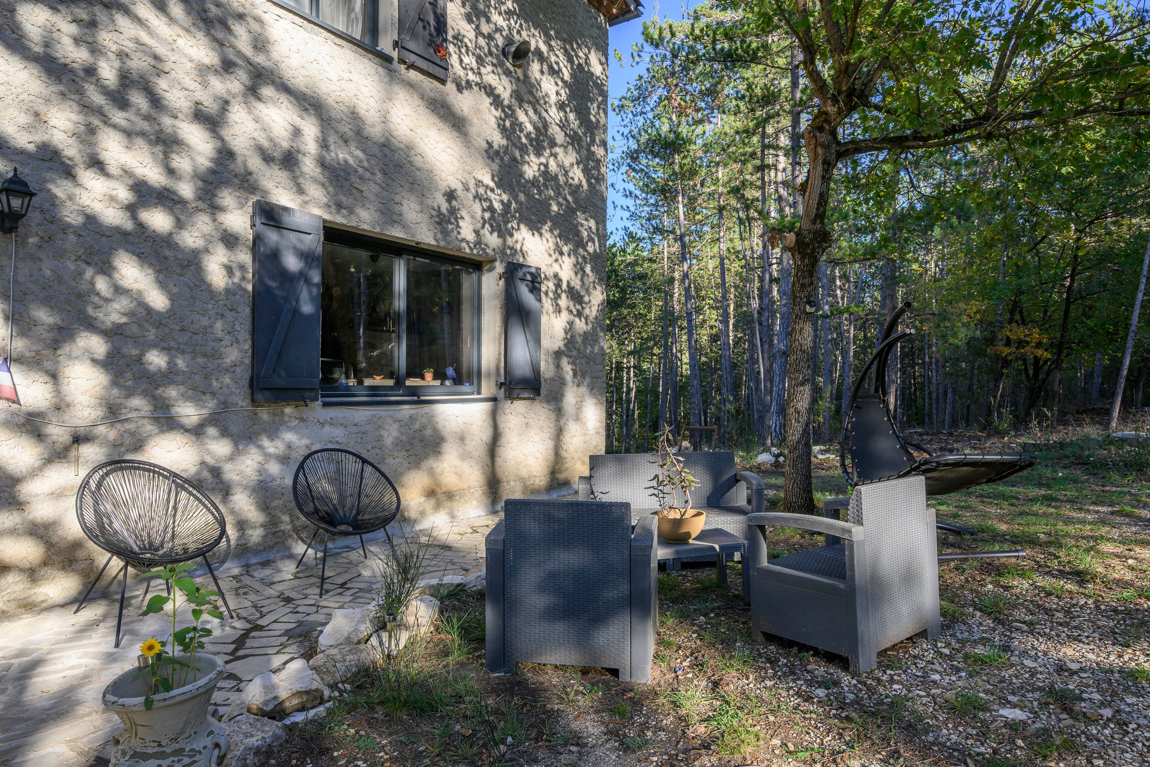 Un edificio de piedra con una ventana, mesas y sillas en la terraza, y un bosque circundante.