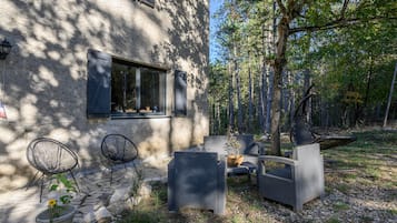 Un edificio de piedra con una ventana, mesas y sillas en la terraza, y un bosque circundante.