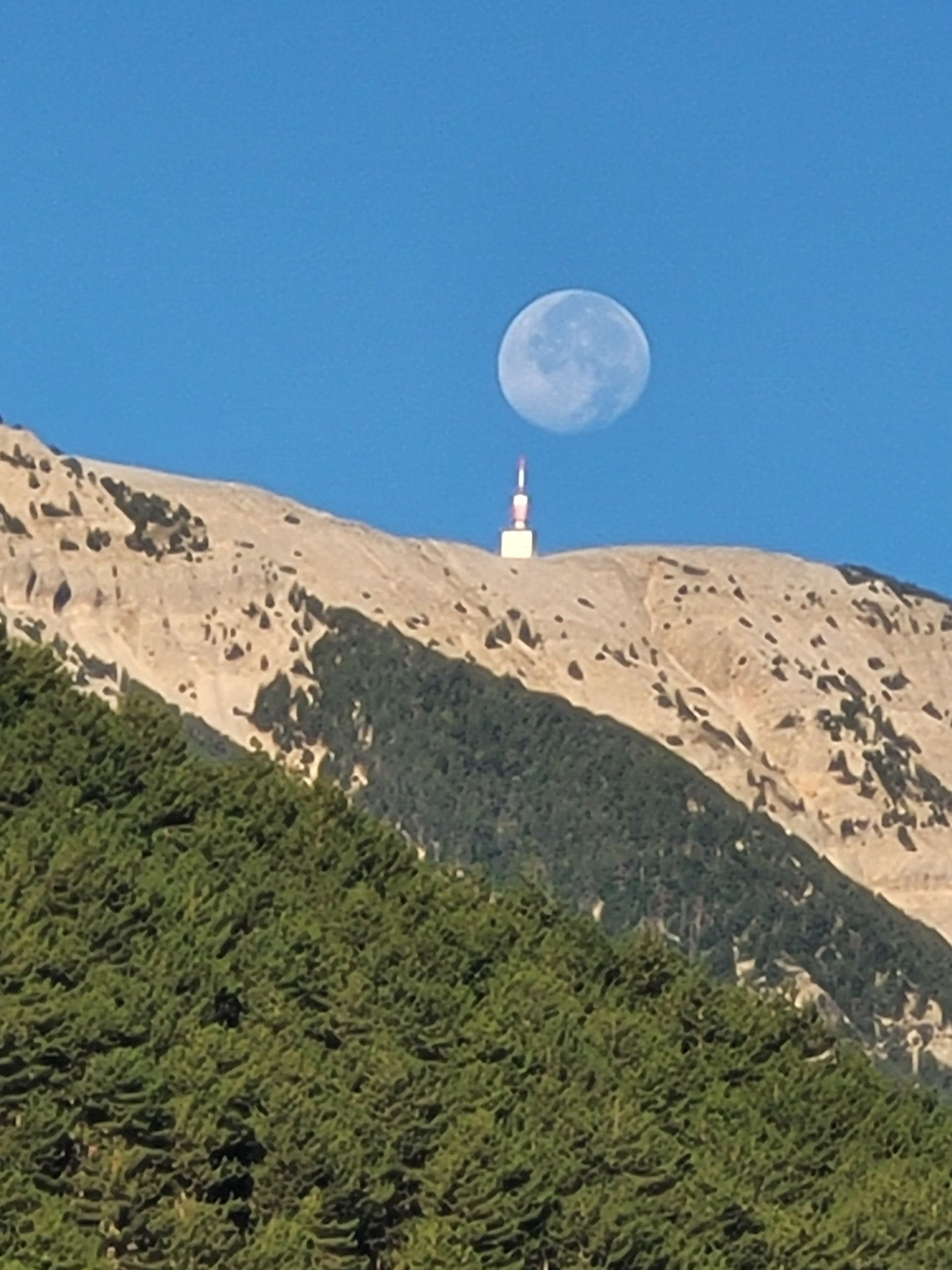 A mountain with a small white structure on top, a clear blue sky, and a large moon in the background.