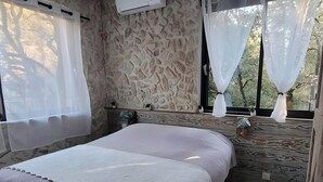 A bedroom with a stone wall, a bed with a white and purple bedspread, an air conditioning unit, and sheer curtains. - La Tour’terelle Guest Room: Mountain View, Private Terrace & Wi-Fi (Brantes)