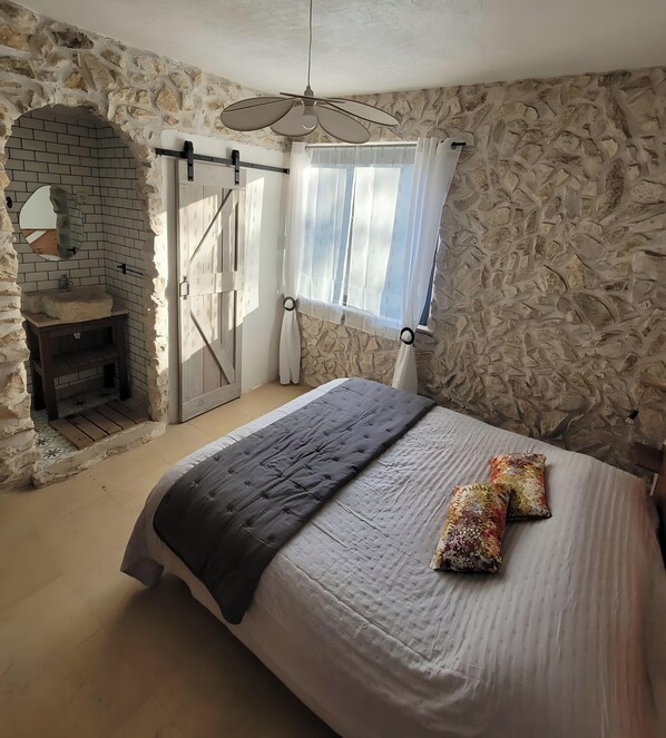 Un dormitorio con pared de piedra, cama con colcha y almohadas, ventana con cortinas y baño con lavabo y espejo.