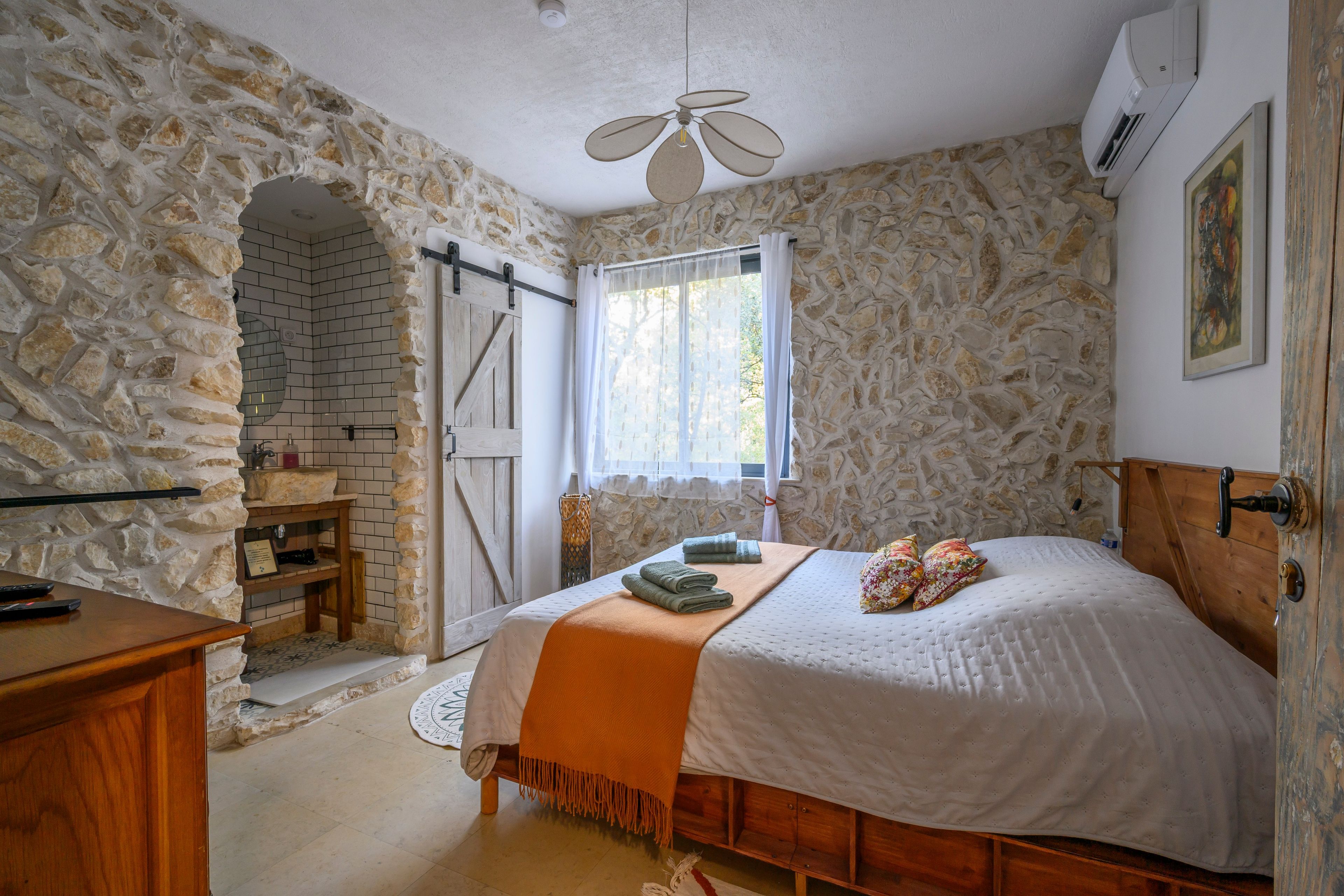 Un dormitorio con pared de piedra, cama con edredón blanco, cabecero de madera y ventilador de techo.