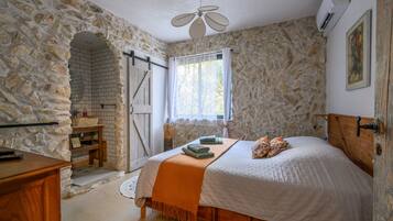 Un dormitorio con pared de piedra, cama con edredón blanco, cabecero de madera y ventilador de techo.