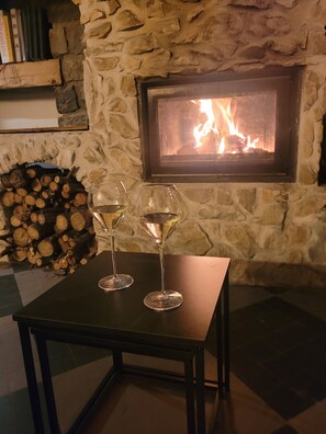 A stone fireplace with a fire, two wine glasses on a table, and a stack of firewood. - L'Estive Guest Room with Mountain View, Wi-Fi, and Air Conditioning (Brantes)