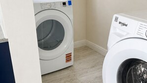 A laundry room with two white Whirlpool washers, a window with a view of a white fence, and a light-colored tiled floor. - Clifton House -Falmouth, Antigua - 3 Bed 2 Bath (St. Paul's)