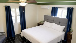 A bedroom with a bed, curtains, a ceiling fan, an air conditioning unit, and a small plant on the nightstand. - Clifton House -Falmouth, Antigua - 3 Bed 2 Bath (St. Paul's)