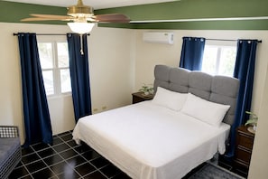 A bedroom with a bed, curtains, a ceiling fan, an air conditioning unit, and a small plant on the nightstand.