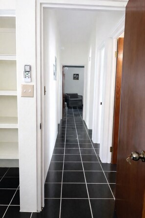 Un pasillo con piso de baldosas blanco y negro, una puerta blanca a la derecha y una silla gris al fondo.