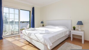 A hotel room with a large bed, a small bedside table with a lamp, a view through a sliding glass door, and a white rug. - B85 - Seasun Step Apartment in Praia da Rocha (Portimão)