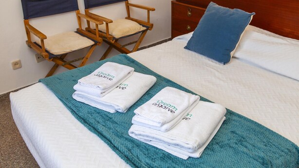 A neatly made bed with folded towels and a teal blanket, wooden chairs, a dresser, and a blue pillow. - A75 - Casa da Ponte 1 Bed Flat in Lagos (Lagos)