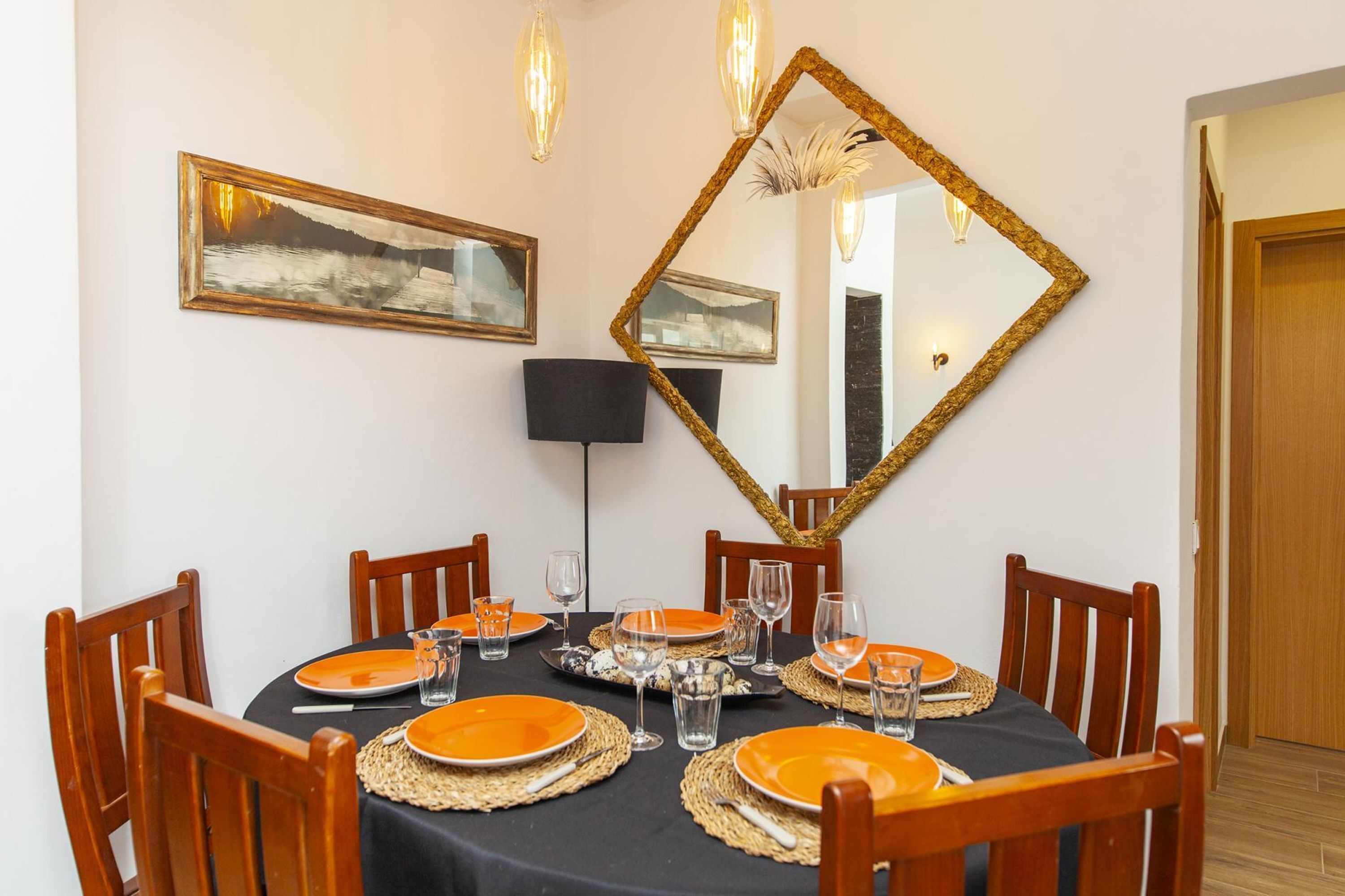 Un comedor con una mesa redonda dispuesta para seis personas, rodeada de sillas de madera. Hay cuadros enmarcados en la pared, un espejo grande y una lámpara de pie.