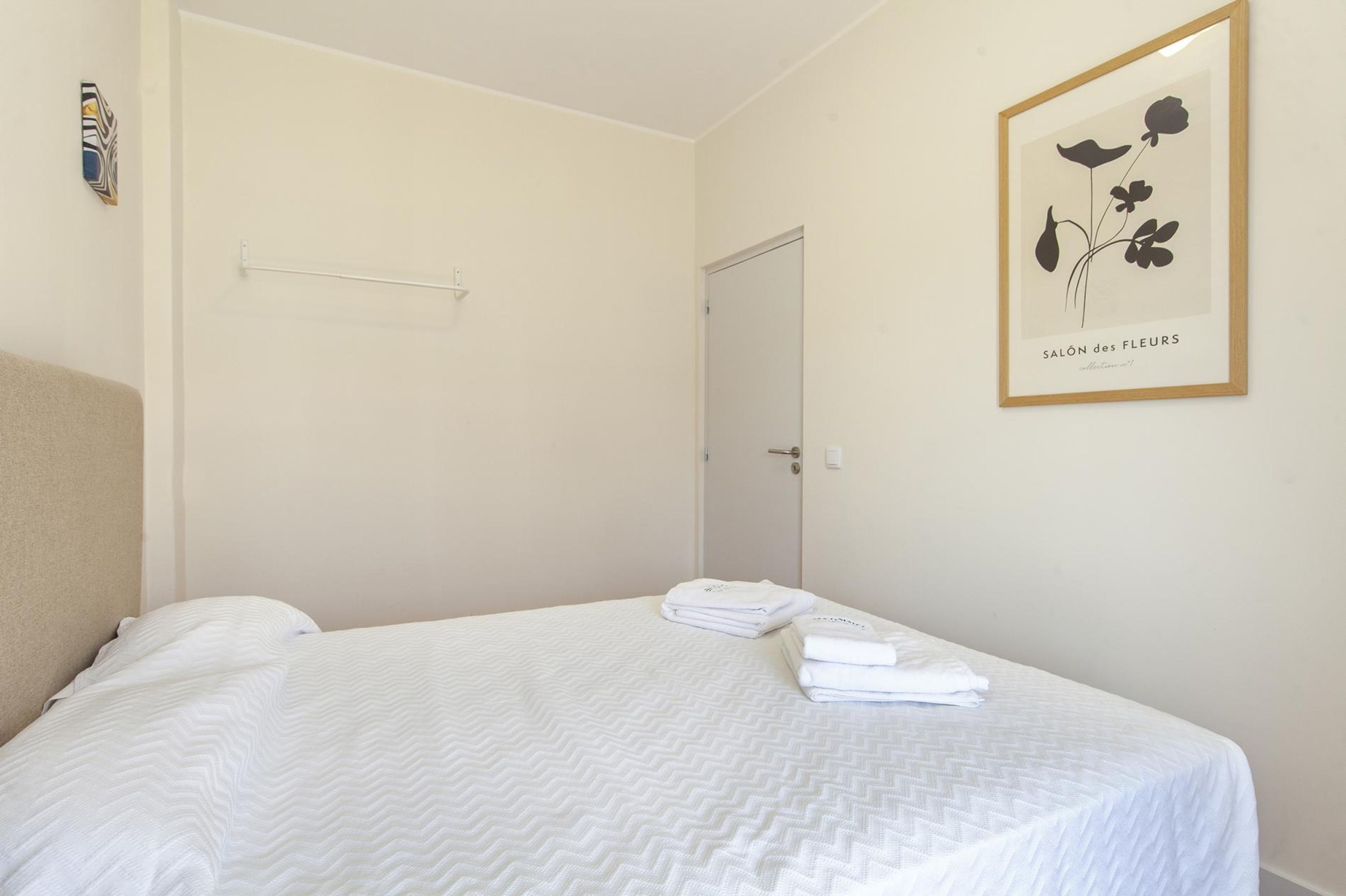 A neatly made bed with white linens and folded towels, a white door, and a framed botanical print on the wall.