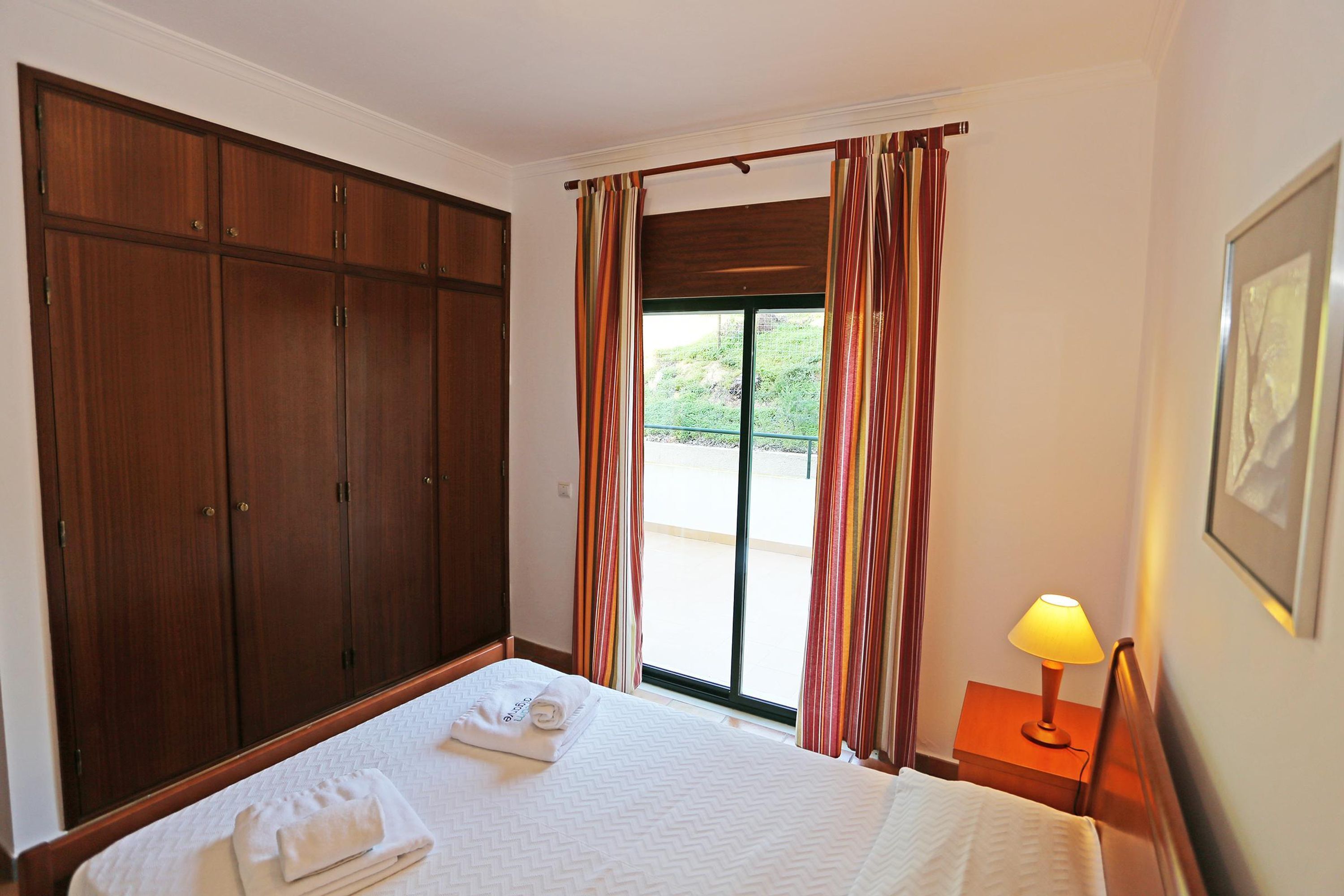 A hotel room with a bed, a wooden wardrobe, a window with curtains, and a nightstand with a lamp.