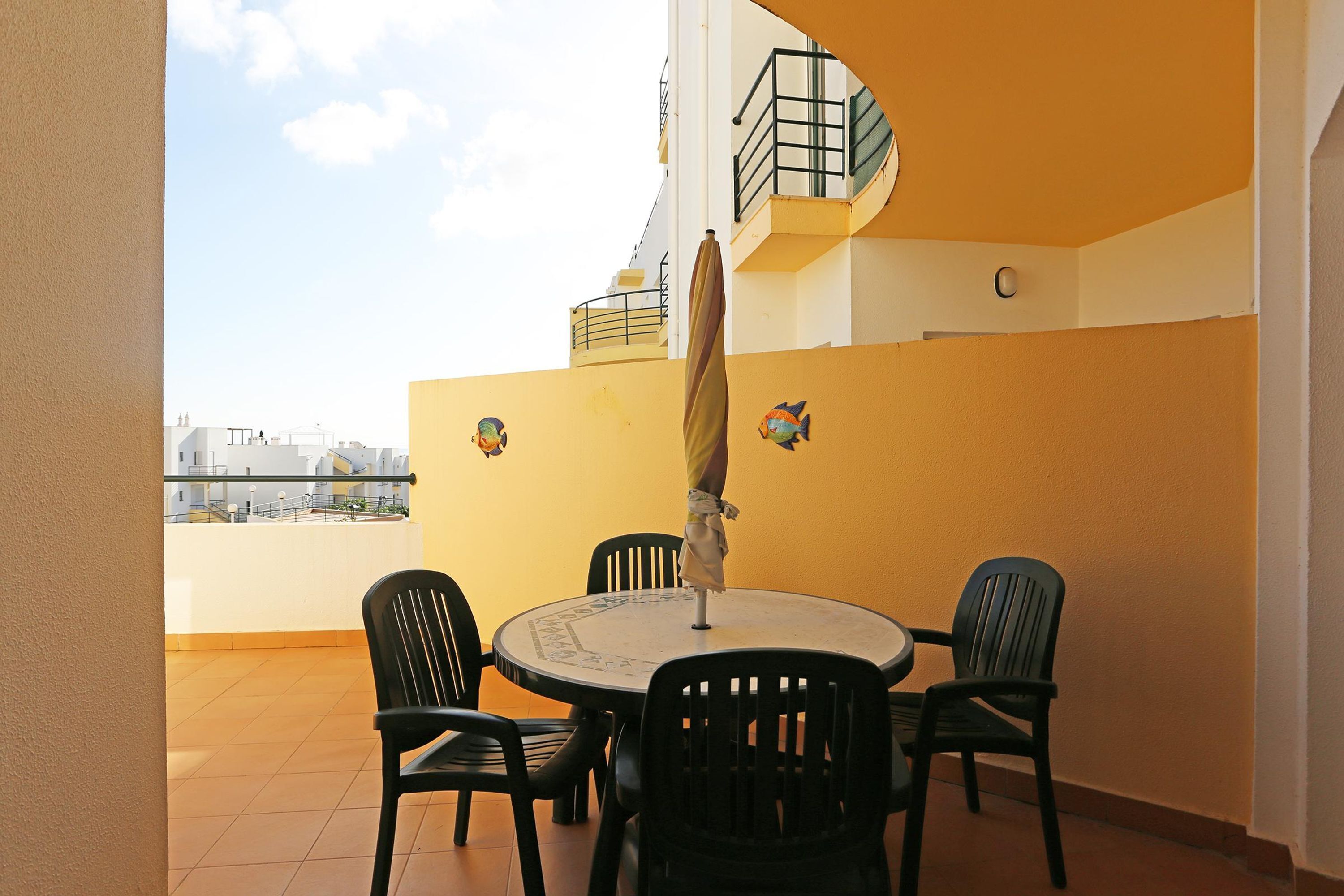 Een ronde tafel met vier stoelen en een parasol, geplaatst op een balkon met gele muren en uitzicht op gebouwen.