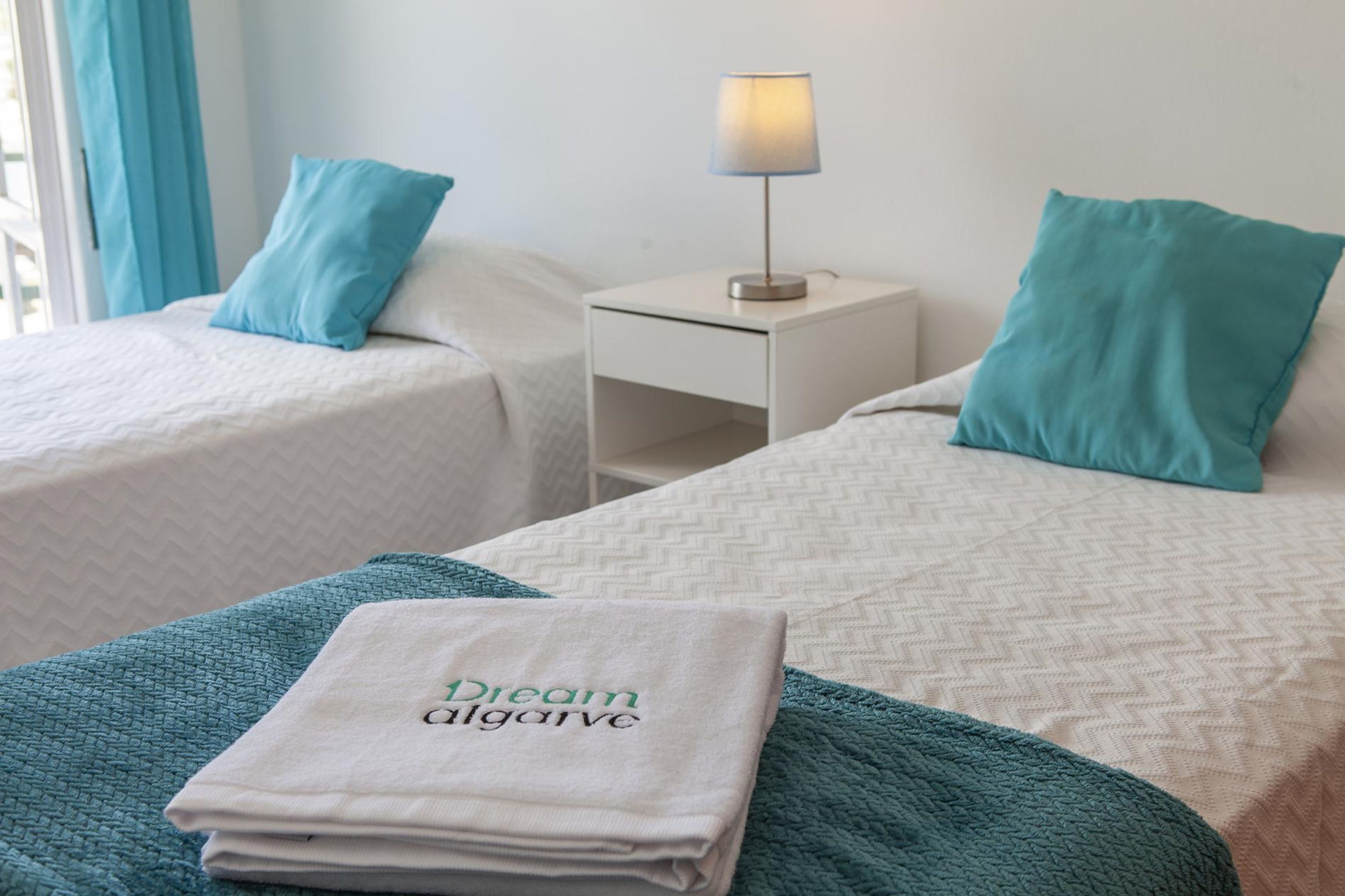 A hotel room with two beds, each with a teal pillow and a white blanket. There is a nightstand with a lamp between the beds. A folded towel with "Dream Algorve" text is on the bed.