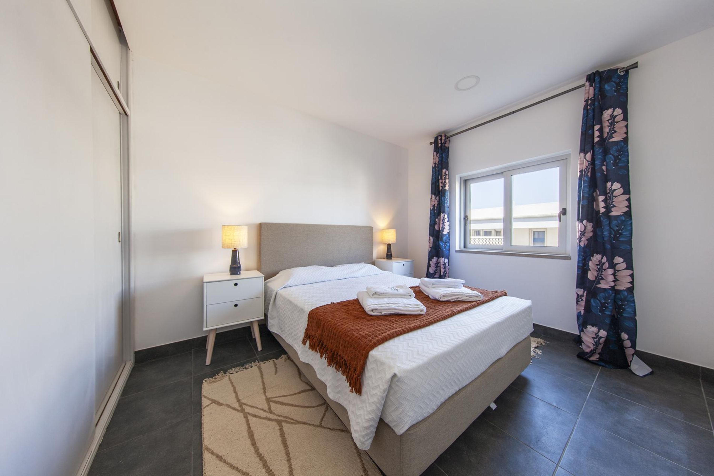 Una camera da letto con un letto, comodini, una finestra con tende e vista sugli edifici.