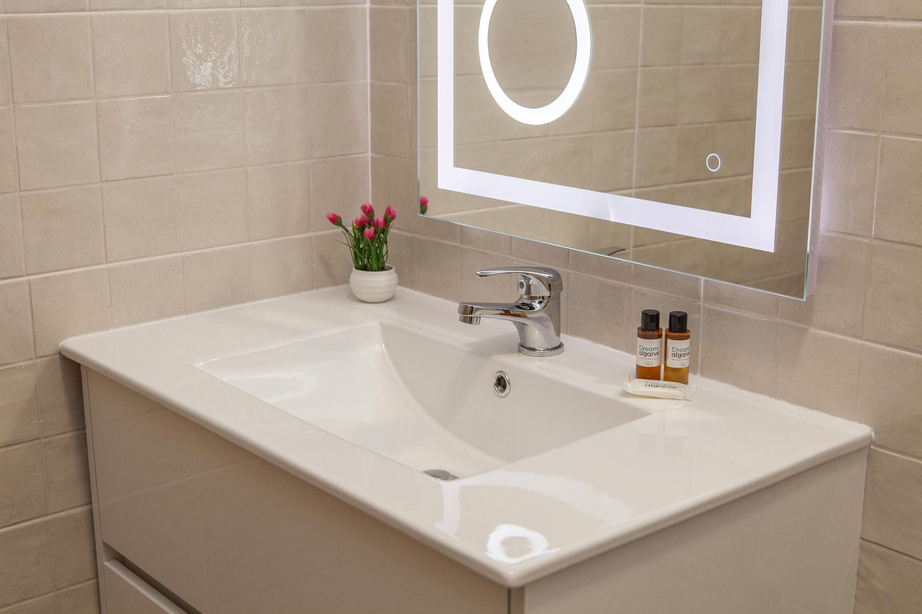 Ein Badezimmer mit weißem Waschbecken, einem Spiegel mit runder Beleuchtung und einem kleinen Blumentopf mit rosa Blüten.
