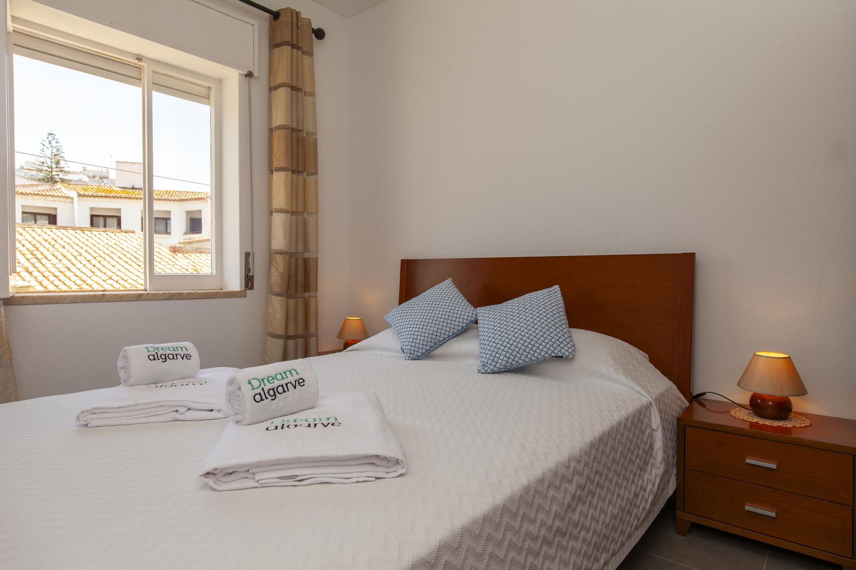 A neatly made bed with two pillows, a bedside lamp, and a nightstand with a lamp. There are folded towels on the bed and a sign with the hotel name.