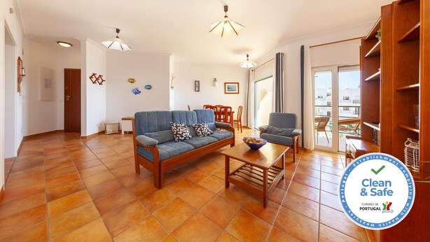 Amplia sala de estar con sofá, mesa de centro y sillas. Se aprecia una estantería de madera y una puerta que conduce a otra habitación.