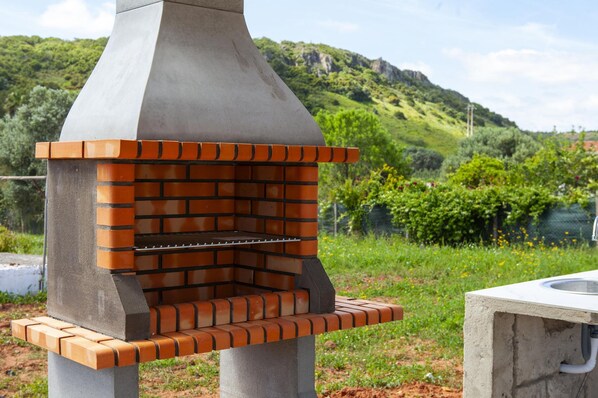 A brick-built outdoor grill with a chimney, set against a lush green hillside.