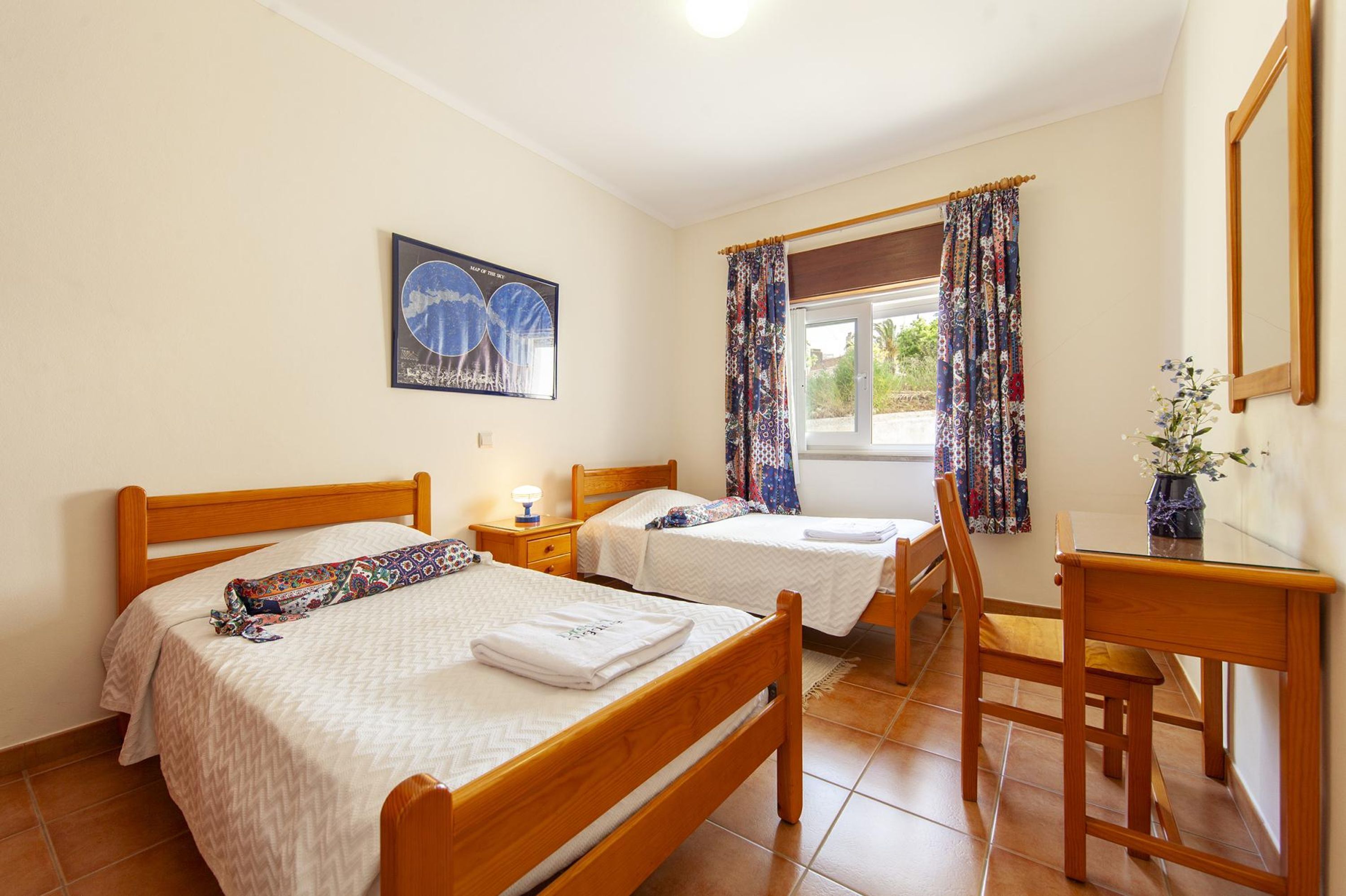 A hotel room with two beds, a desk, a chair, a window with curtains, and a framed picture on the wall.